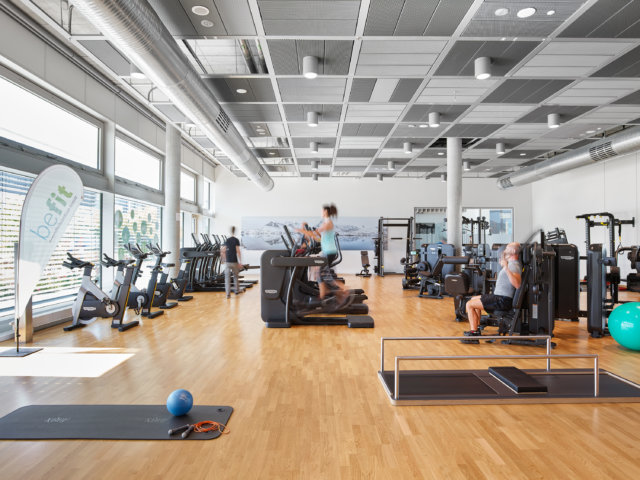 Innenansicht eines Fitnessstudioraumes. Mehrere Menschen trainieren an verschiedenen Geräten.